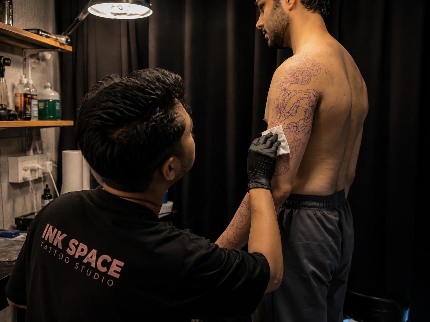 A tattoo artist carefully wiping and checking a purple stencil applied to a client’s upper arm before beginning the tattoo.