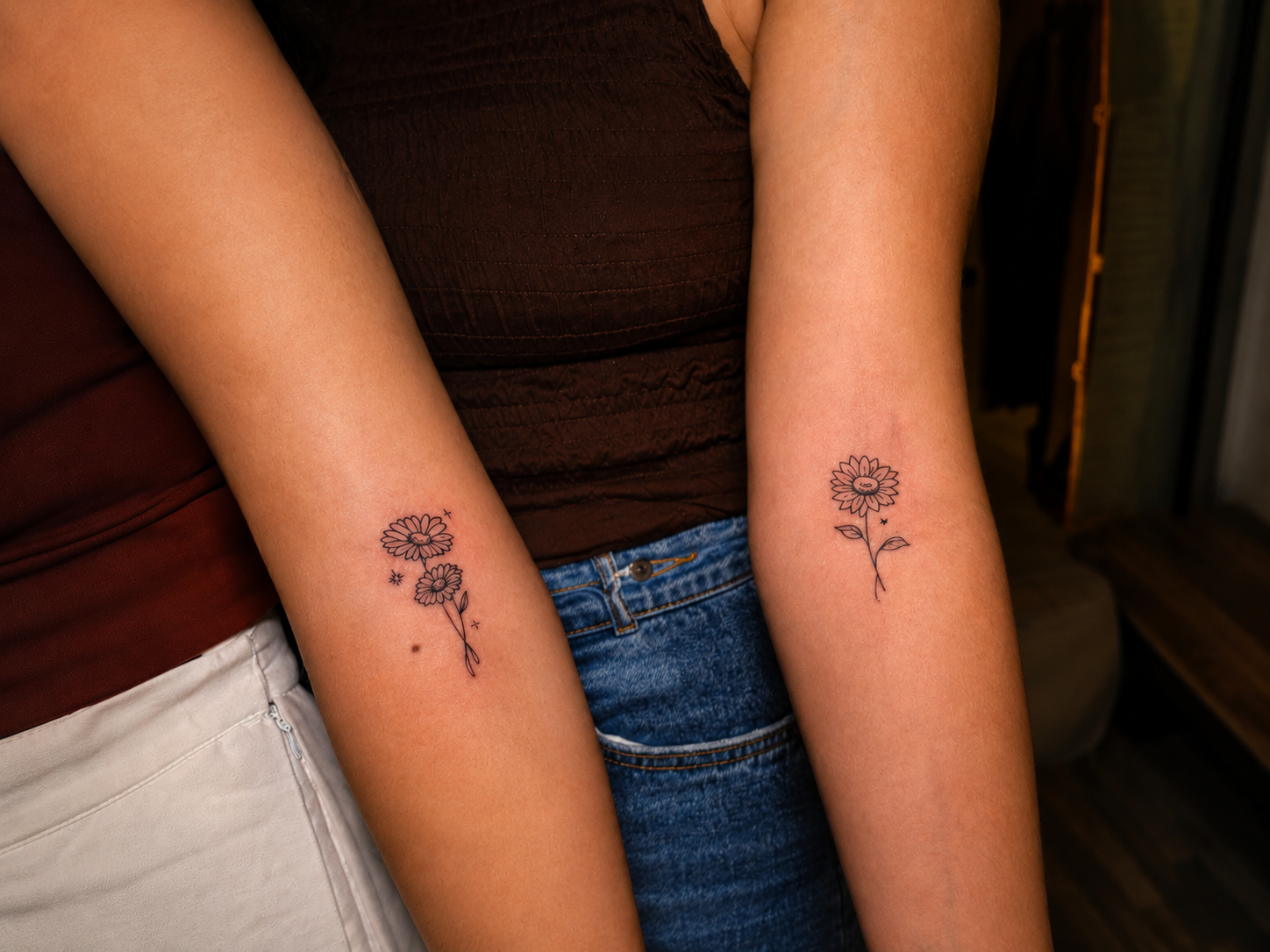 Close-up of matching fine-line floral tattoos on two forearms, showing delicate daisy designs with leaves and small decorative accents.