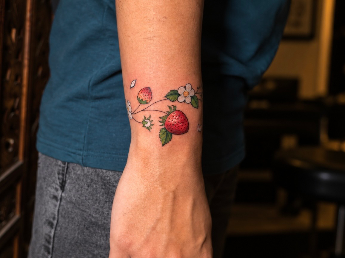 Close-up of a colorful fine-line strawberry and flower tattoo wrapping around the forearm above the wrist.