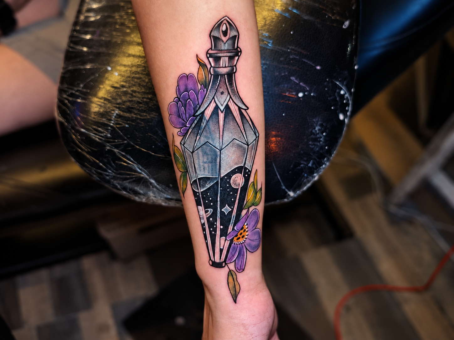 Close-up of a colorful potion bottle tattoo filled with a starry night scene, surrounded by purple flowers and leaves on a forearm.