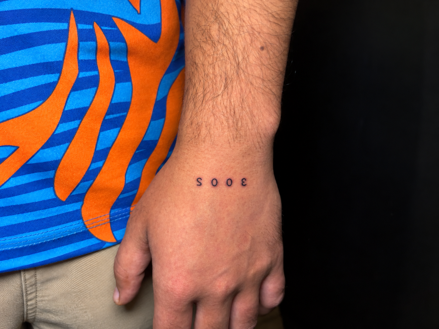 Minimalist numerical tattoo on the wrist of a person wearing a blue and orange patterned shirt, photographed in a close-up.