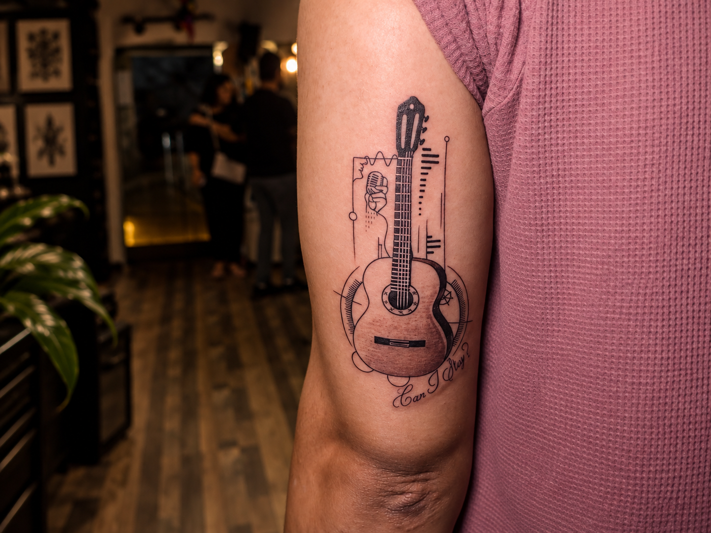 A detailed guitar tattoo on the upper arm, featuring a vintage microphone, geometric linework and the words “Can I Play?” in a warm, softly lit tattoo studio.