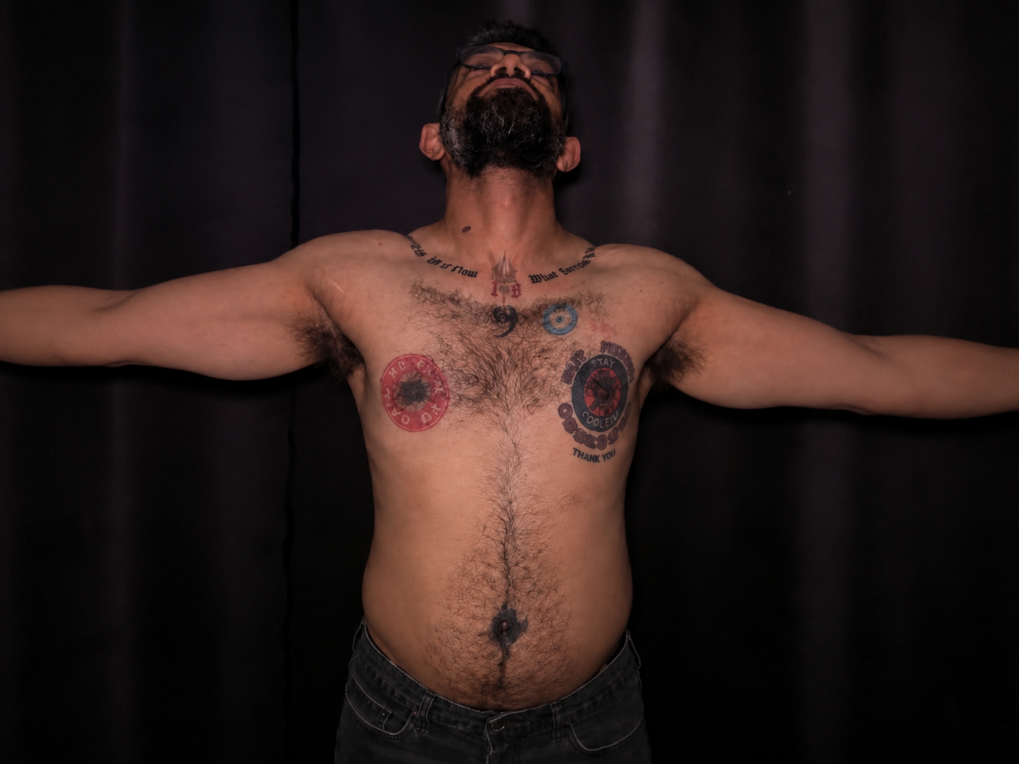 A tattooed man standing with his arms extended in a dark studio, showing multiple tattoos across his chest and shoulders.