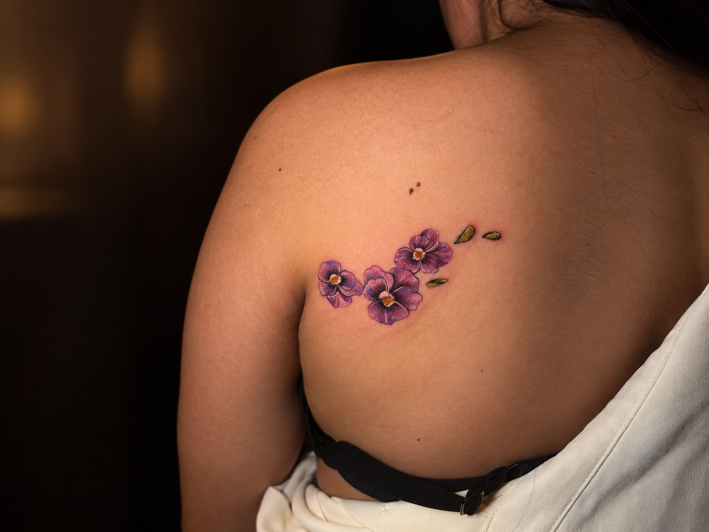 Close-up of a shoulder featuring a delicate purple floral tattoo with small leaves, photographed against a dark studio background.