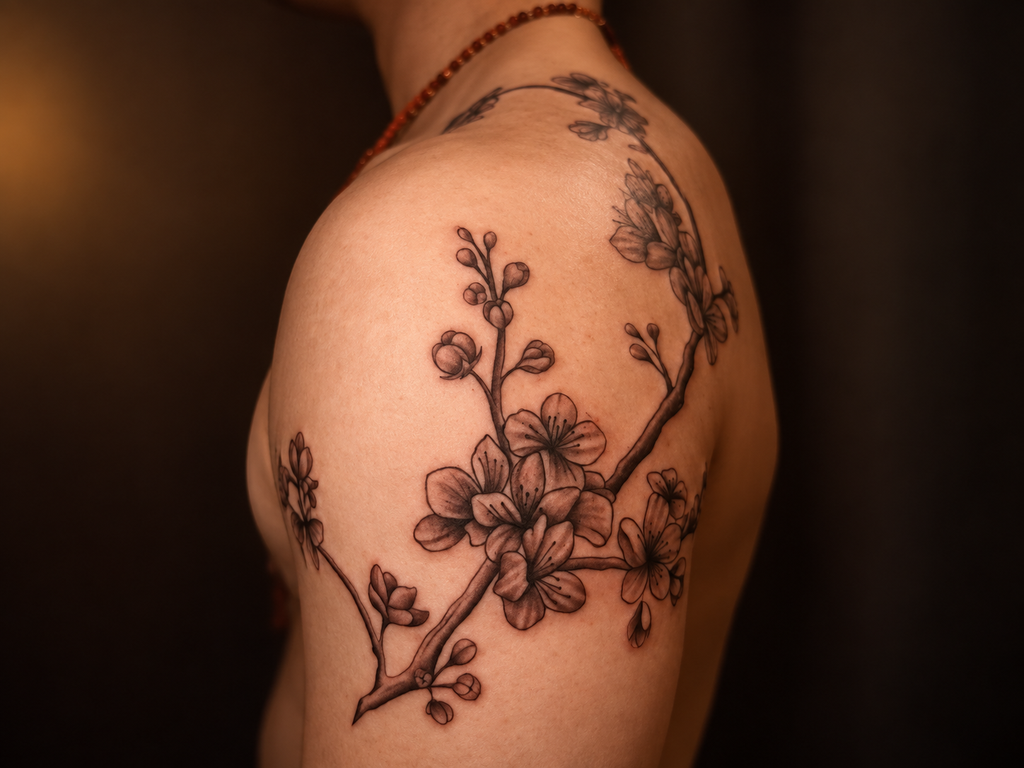 Close-up of a shoulder featuring an ornamental floral tattoo with detailed blossoms and branching stems in warm black and grey tones.