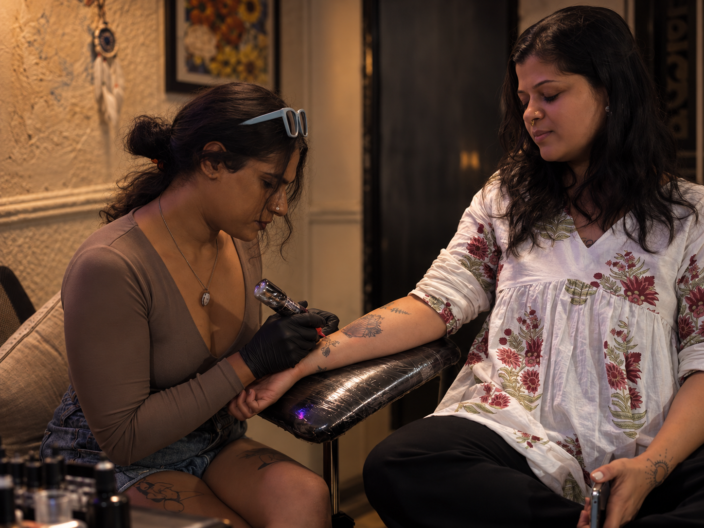 A tattoo artist carefully working on a floral tattoo on a client's forearm inside a warm, intimate tattoo studio.