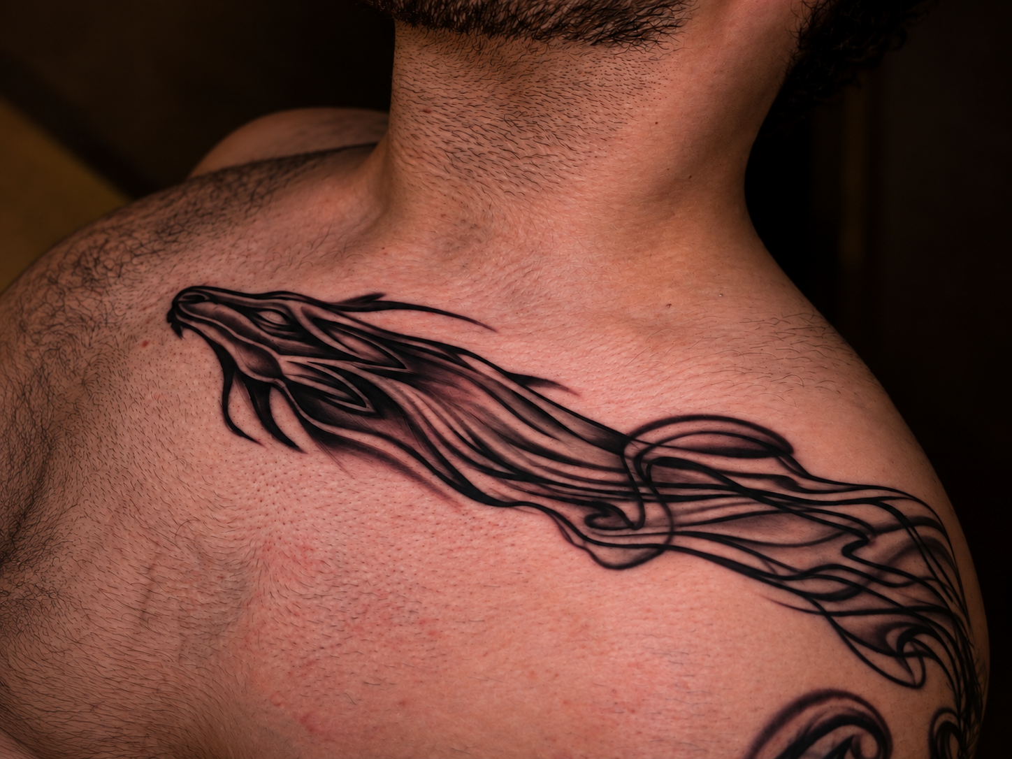 Close-up landscape photograph of a black-and-gray flowing tattoo wrapping across a man's upper chest, collarbone and shoulder, with bold curved lines, layered shading and organic tendrils.