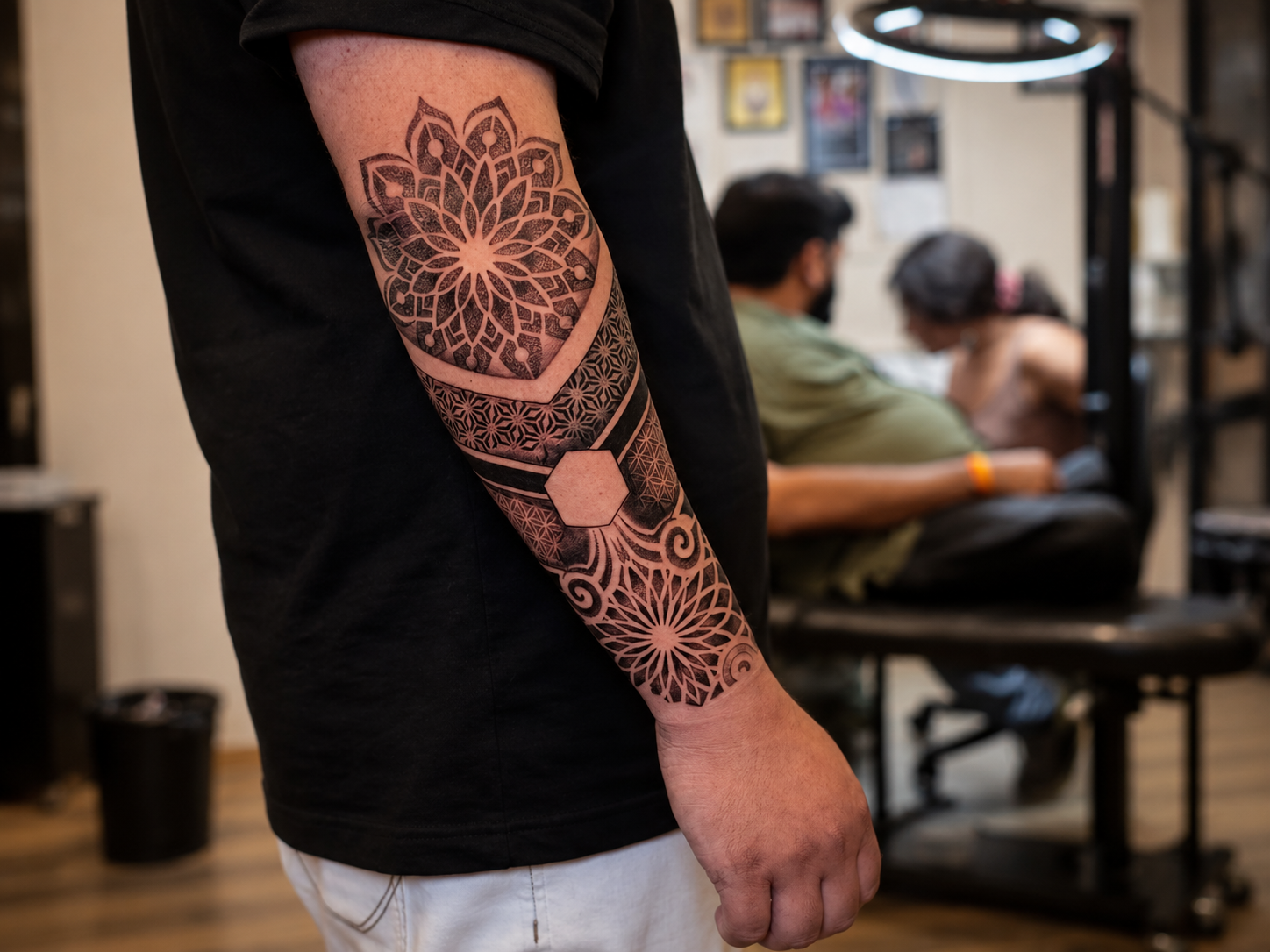 Close-up of a detailed black and grey geometric mandala tattoo covering the arm, featuring lotus-inspired shapes, intricate patterns and geometric bands in a tattoo studio.