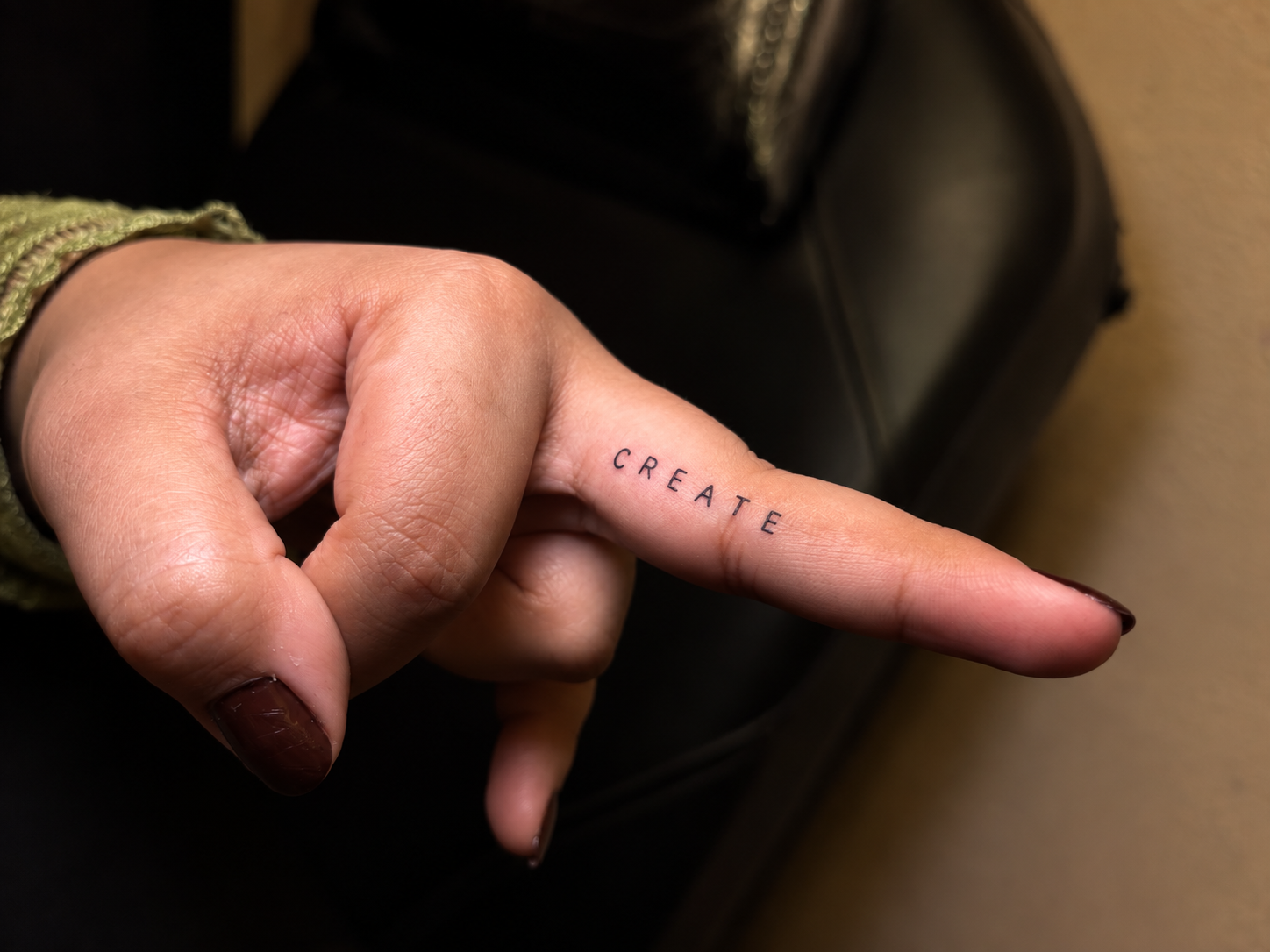 A close-up of a hand featuring a minimalist fine-line “CREATE” tattoo across the index finger.