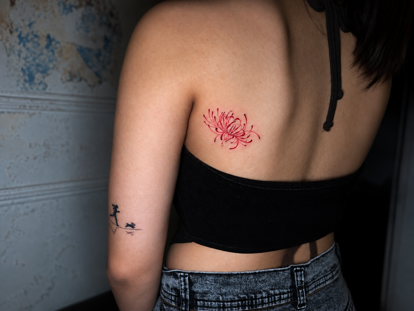 A close-up of a woman’s back showing a delicate red floral tattoo on the upper back and a small black linework tattoo on the upper arm.