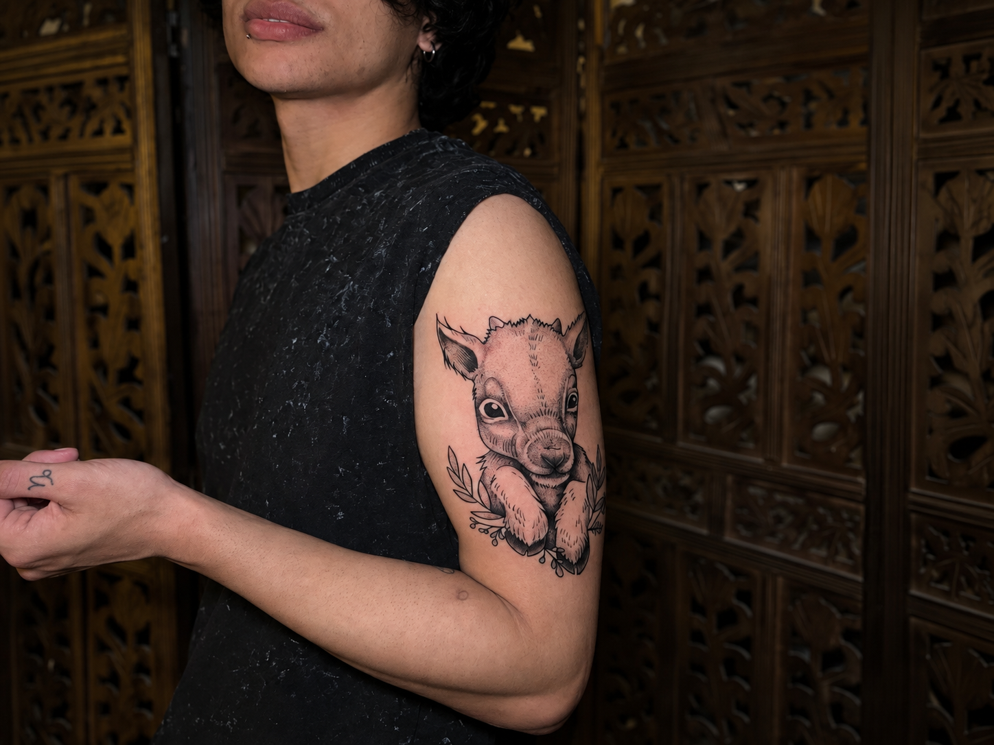 A person showing a detailed baby animal tattoo on the upper arm, photographed in a tattoo studio with a carved wooden backdrop.