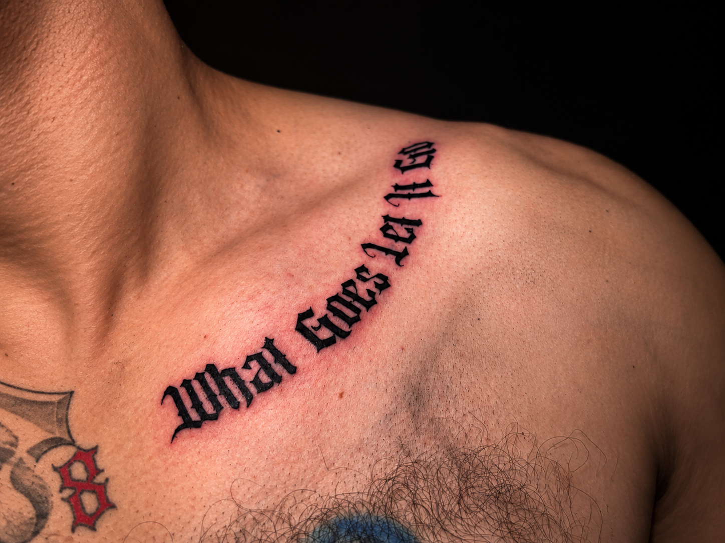 A close-up of a black Gothic lettering tattoo reading “What Will Be” across the collarbone and upper shoulder.
