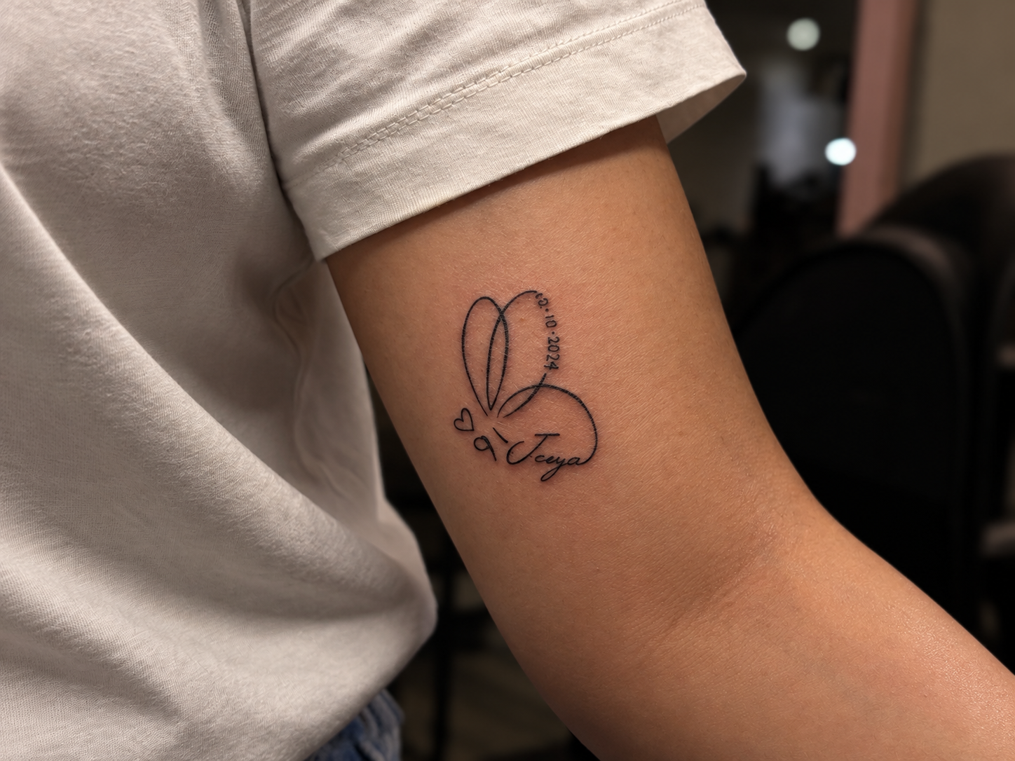 Close-up photograph of a fine-line tattoo on the upper arm, featuring two overlapping hearts, a small date, and a handwritten name, symbolizing a personal memory.