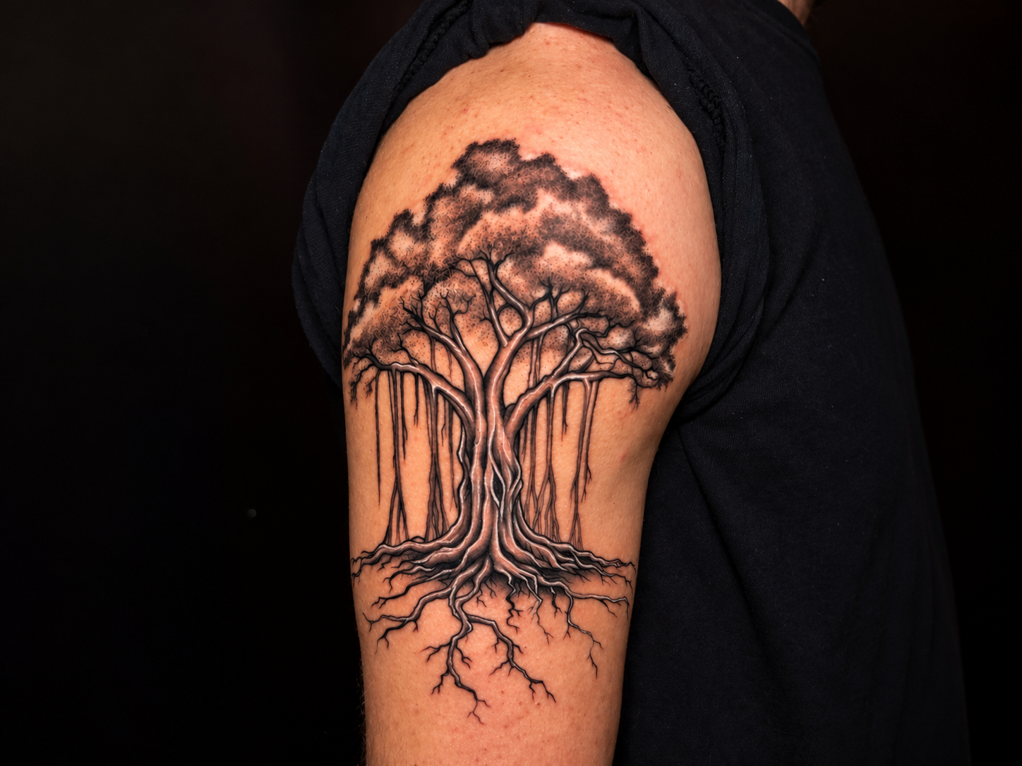 Close-up of a detailed black and grey tree tattoo on the upper arm, showing a textured canopy, intricate branches, hanging lines, and spreading roots against a dark background.