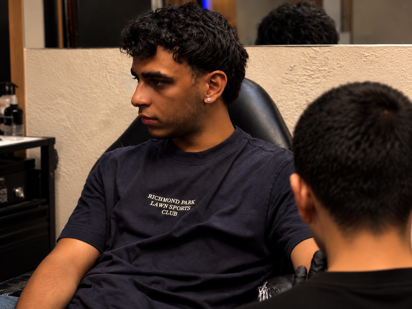 Young man sitting in a tattoo studio chair during a tattoo consultation, with a tattoo artist partially visible in the foreground.